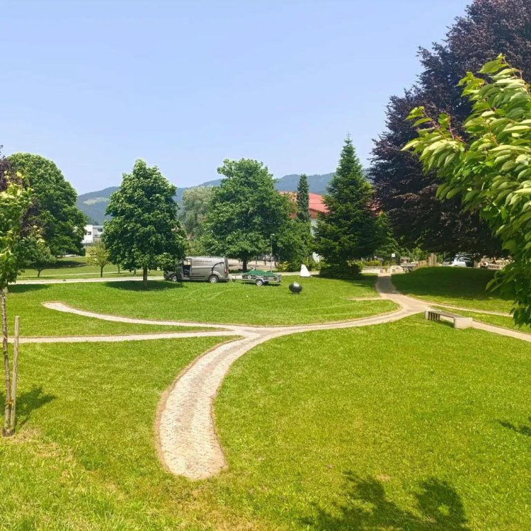 Pokošena parkovna zelenica z drevesi in vozilom Mojstra Jake v ozadju, v bližini Slovenj Gradca