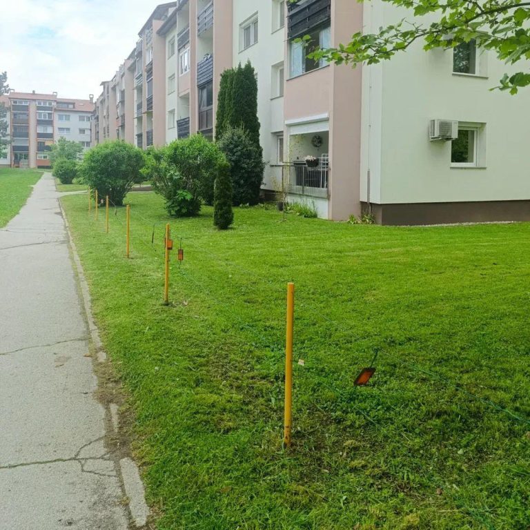 Pokošena zelenica z označevalnimi koli pred večstanovanjskim objektom